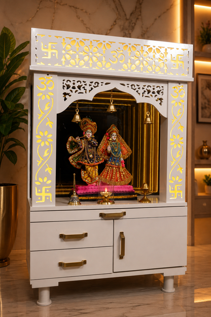A white wooden Hindu home altar (holy cabinet) with gold accents, featuring a carved swastika at the top, a central arched niche with hanging brass bells, and a deity idol inside, set against a warm, modern home interior background.