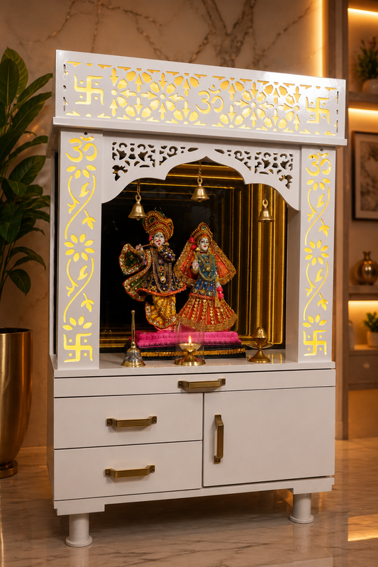 A white wooden Hindu home altar (holy cabinet) with gold accents, featuring a carved swastika at the top, a central arched niche with hanging brass bells, and a deity idol inside, set against a warm, modern home interior background.