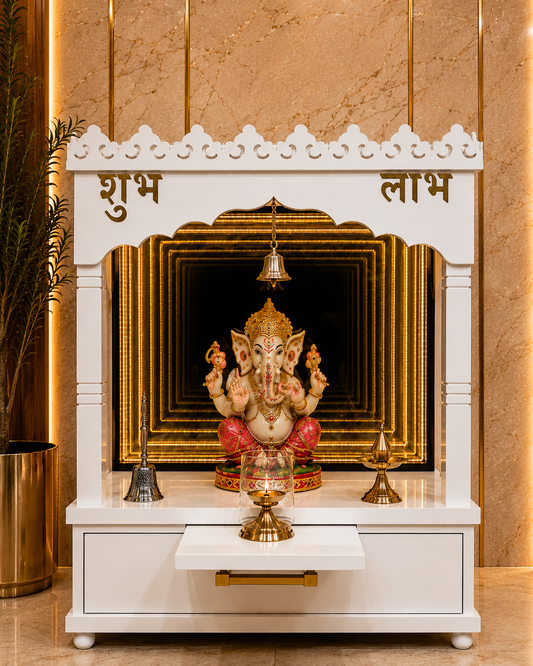 A white and gold Ganesha idol housed in a decorative white and gold temple-style cabinet with brass bells and a glass candle holder, set against a beige marble wall with vertical gold accents.