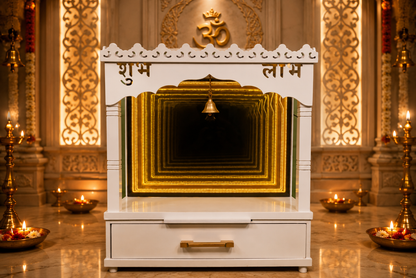 A white wooden Shubh Lamk temple altar with gold accents, featuring a carved Om symbol on the top arch, a hanging brass bell, and a drawer with a gold handle, set against a decorated temple backdrop with oil lamps.