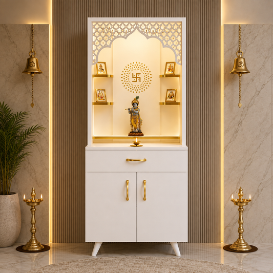 A white and gold Hindu home altar cabinet with a carved arched top panel, built-in shelves, and brass-tone handles and pulls, shown in a styled interior setting with lamps, a plant, and marble walls.